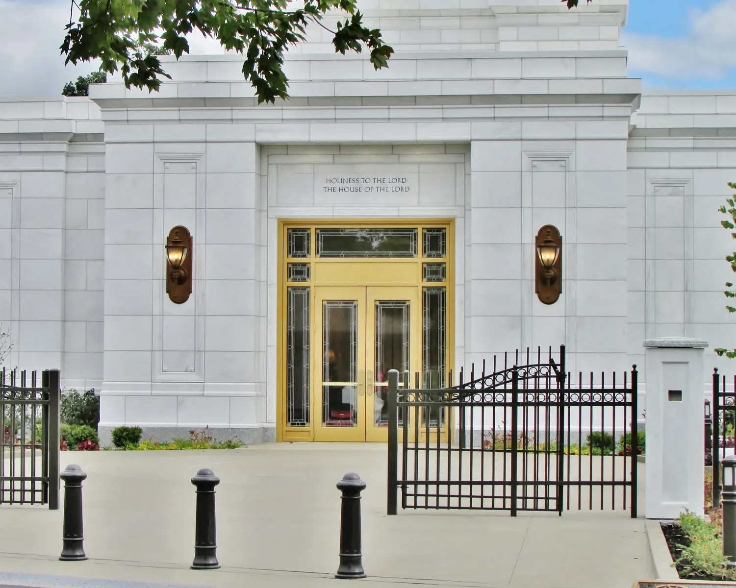 Gold temple entrance doors beneath an engraved inscription, with bronze wall lanterns and a black iron gate in front.