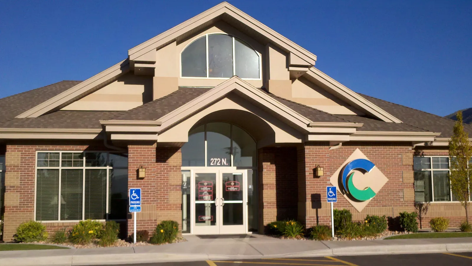Close view of a UCCU branch entry with an arched window, brick columns, tan trim and a diamond-shaped logo sign.