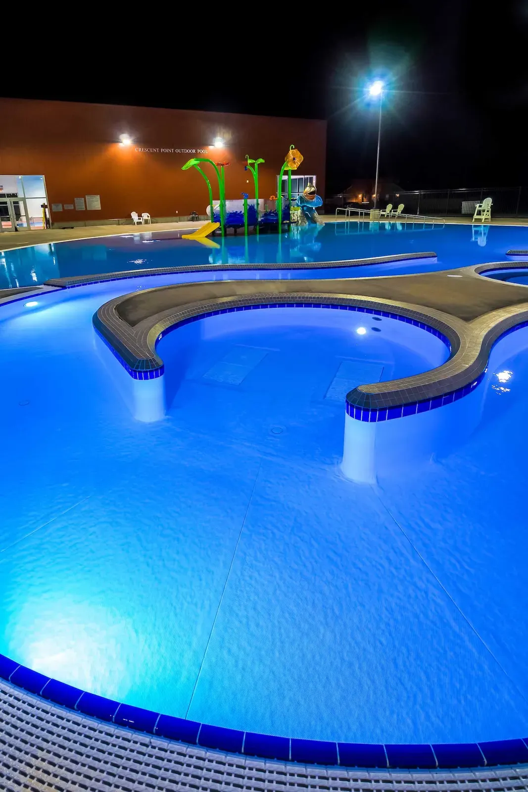 Outdoor leisure pool lit blue at night, with a curved seating ledge and a water play structure beyond