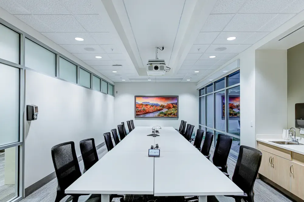 Conference room with a long white table, black chairs and a wall-mounted screen.
