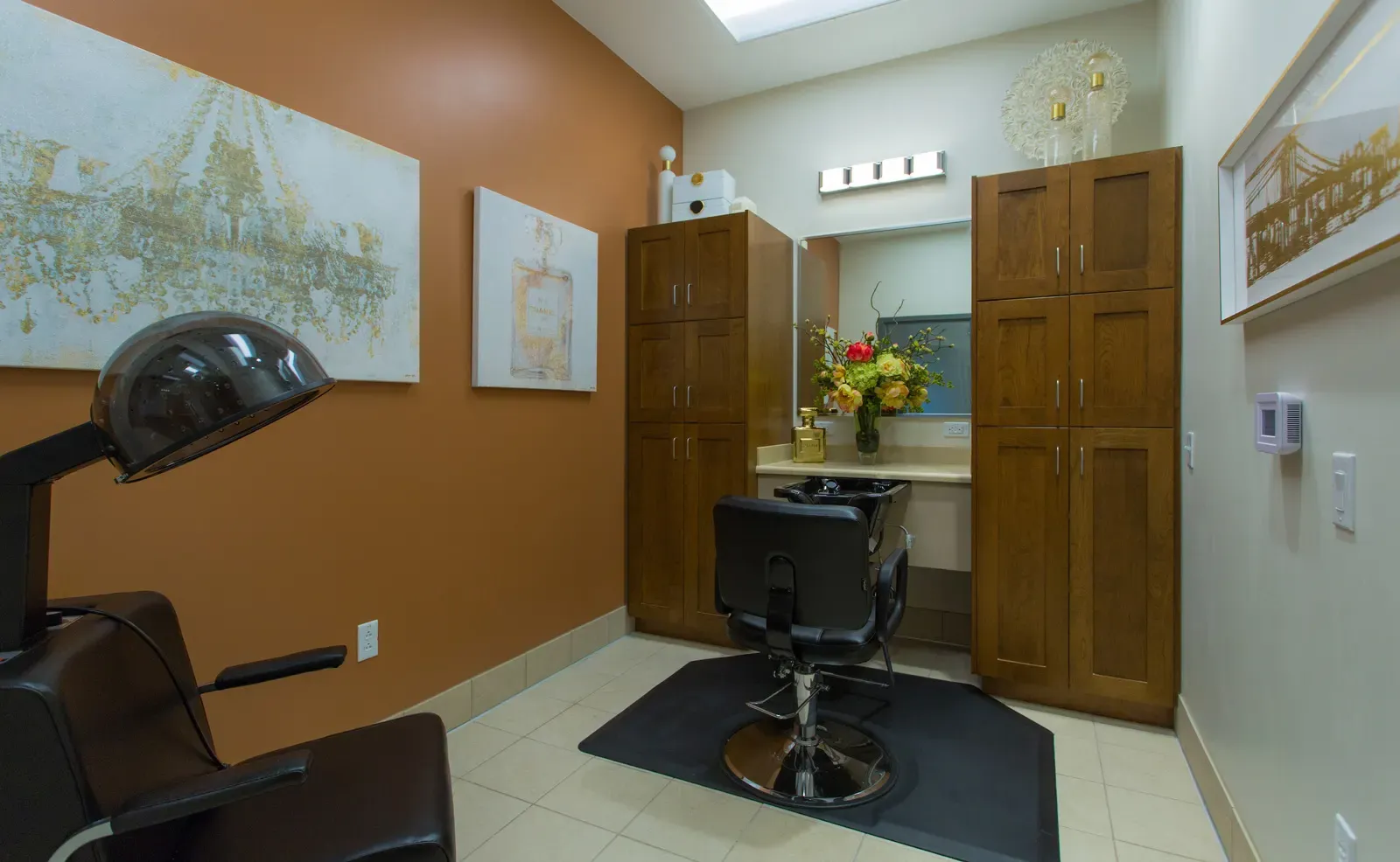 Salon room with a hair-drying chair, a styling chair, and wood cabinets against orange walls.