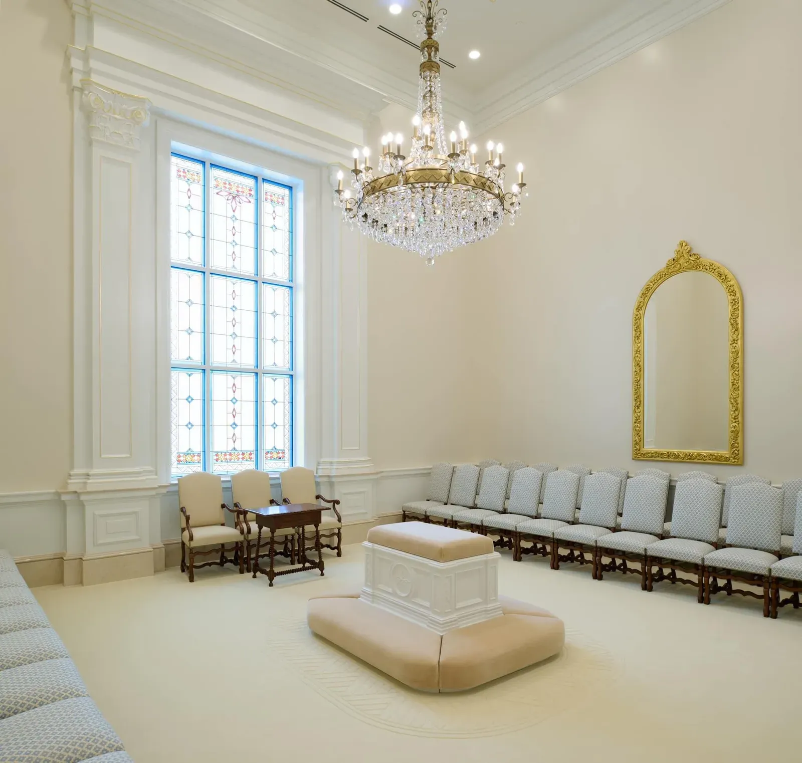 Sealing room with a central cushioned altar, a crystal chandelier, a gold-framed mirror, rows of upholstered chairs and a tall stained-glass window