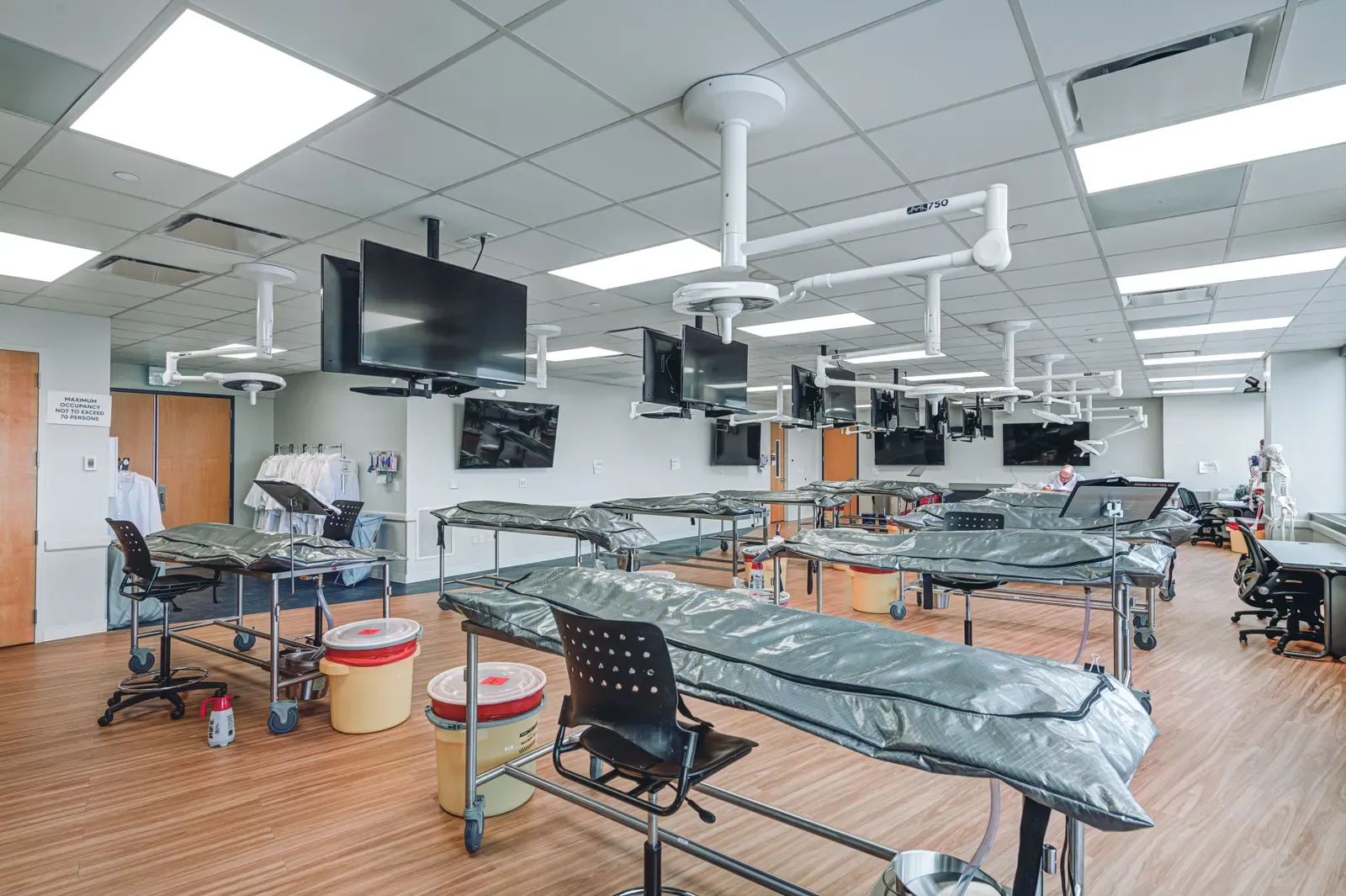 Hands-on teaching lab with covered tables under ceiling-mounted arms and wall monitors, on a wood-look floor.