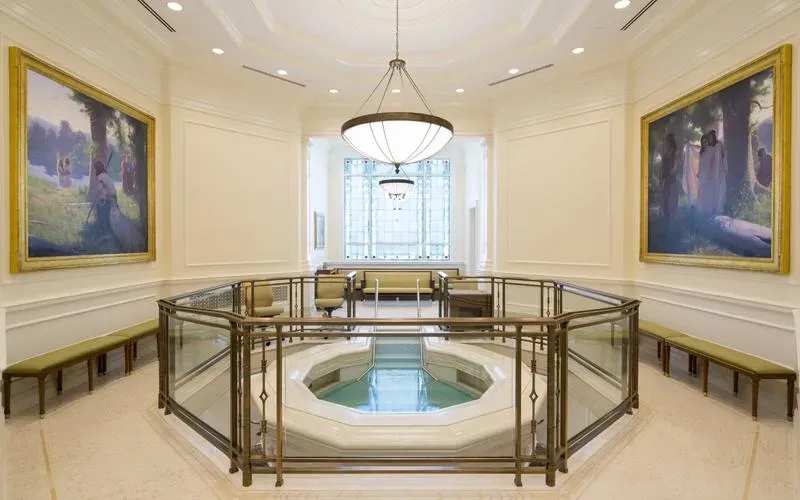 Baptistry with a font behind a glass and brass railing and two large framed paintings on the walls
