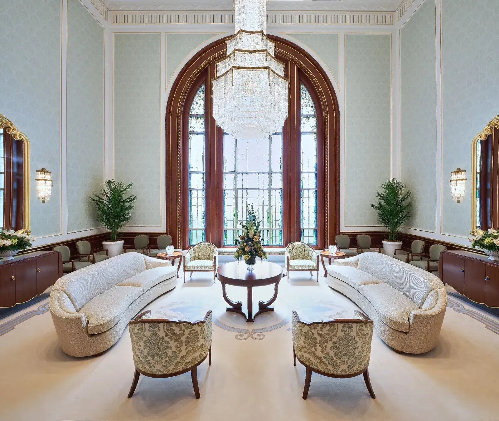 Tall room with a stepped crystal chandelier, two cream sofas, patterned armchairs, a round wood table and arched windows