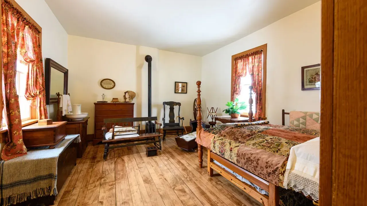 Bedroom with a four-poster bed under a patchwork quilt, a wooden dresser, a small iron stove and floral curtains at the windows.