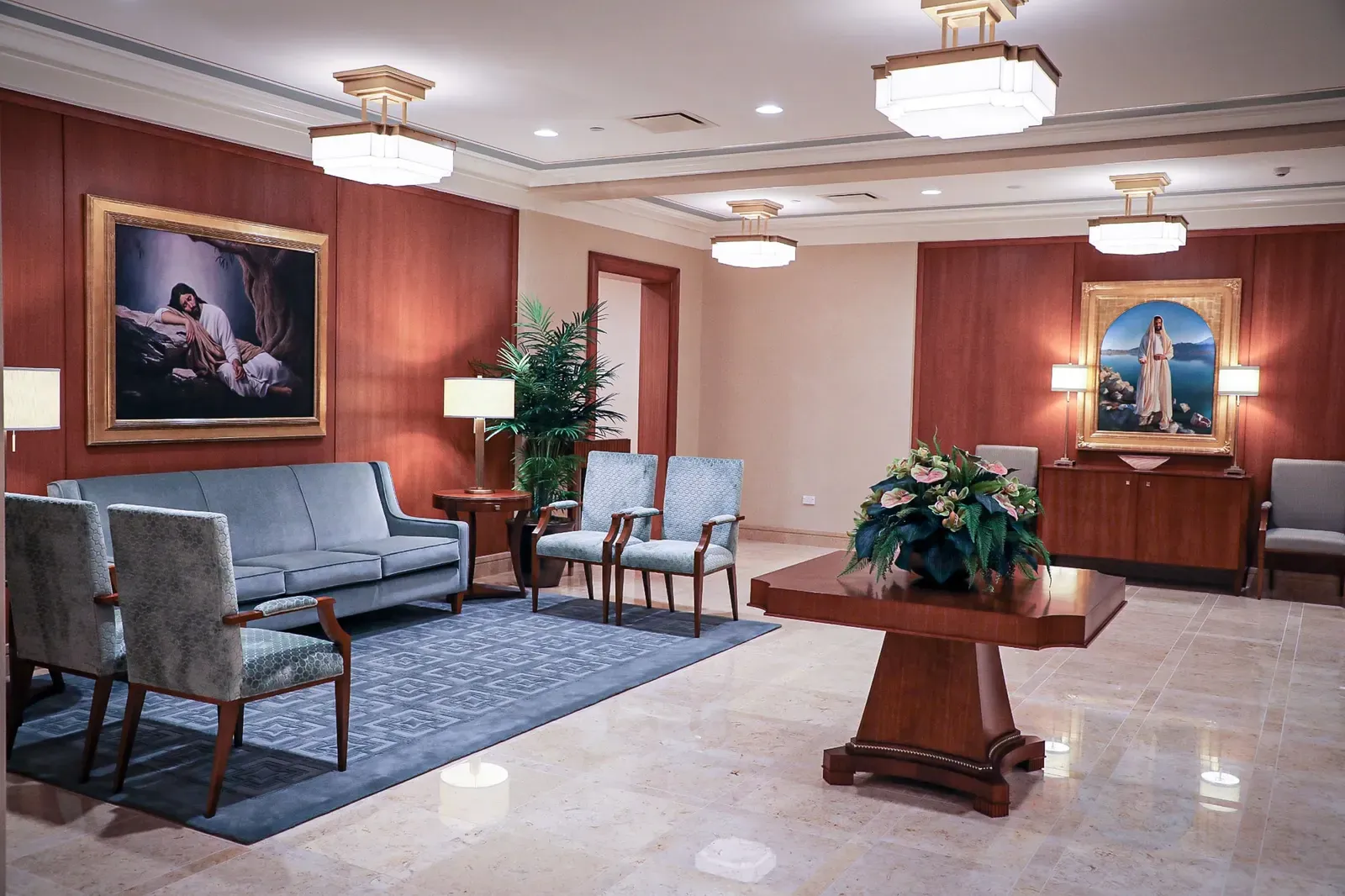 Sitting room with a gray-blue sofa, upholstered armchairs, a polished table with flowers and framed paintings on warm wood paneling