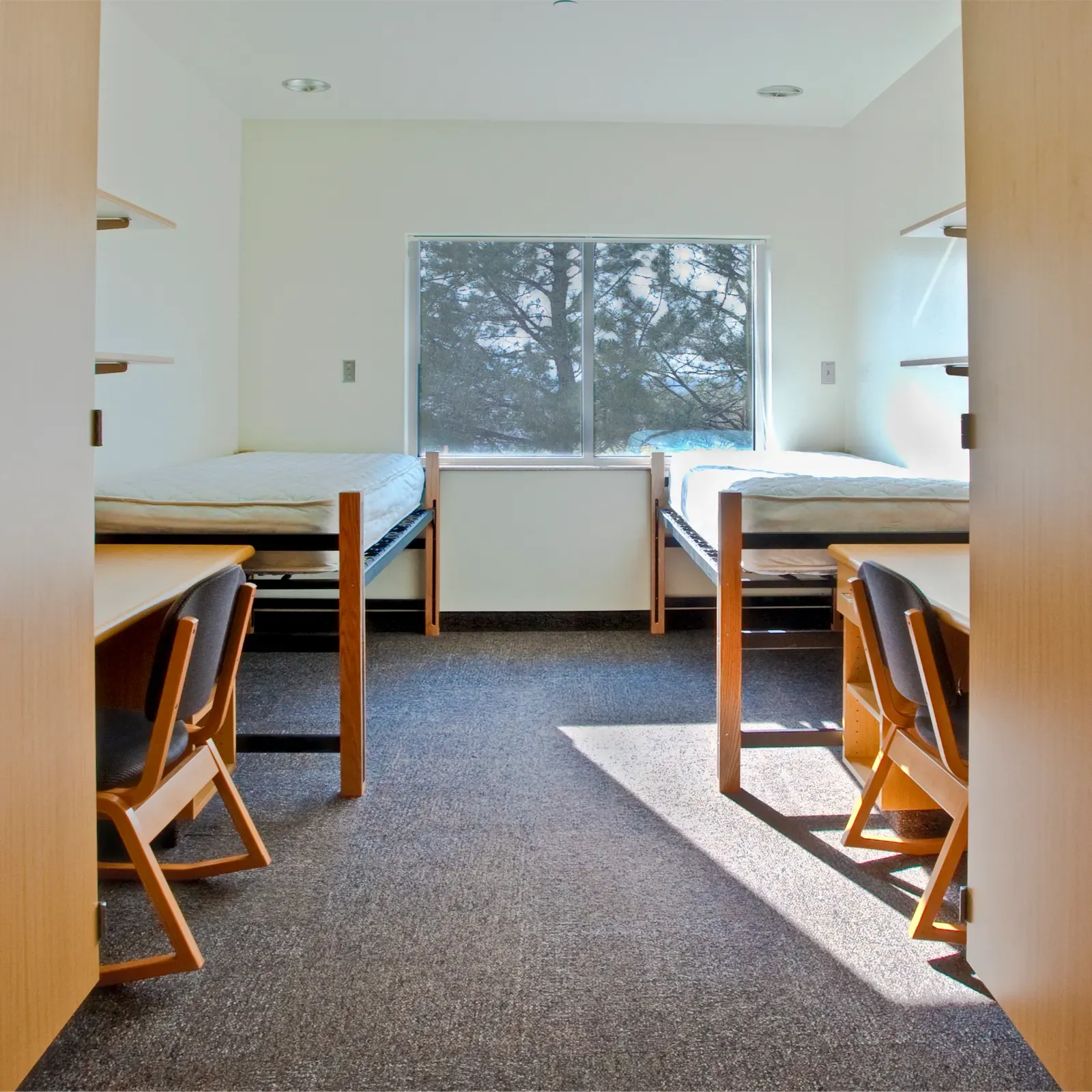 Dorm room with two beds, two desks and a window facing trees.