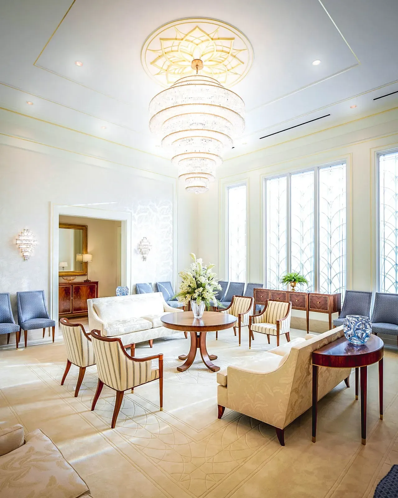 Bright sitting room with a tiered chandelier, tall windows, and cream and blue upholstered seating.
