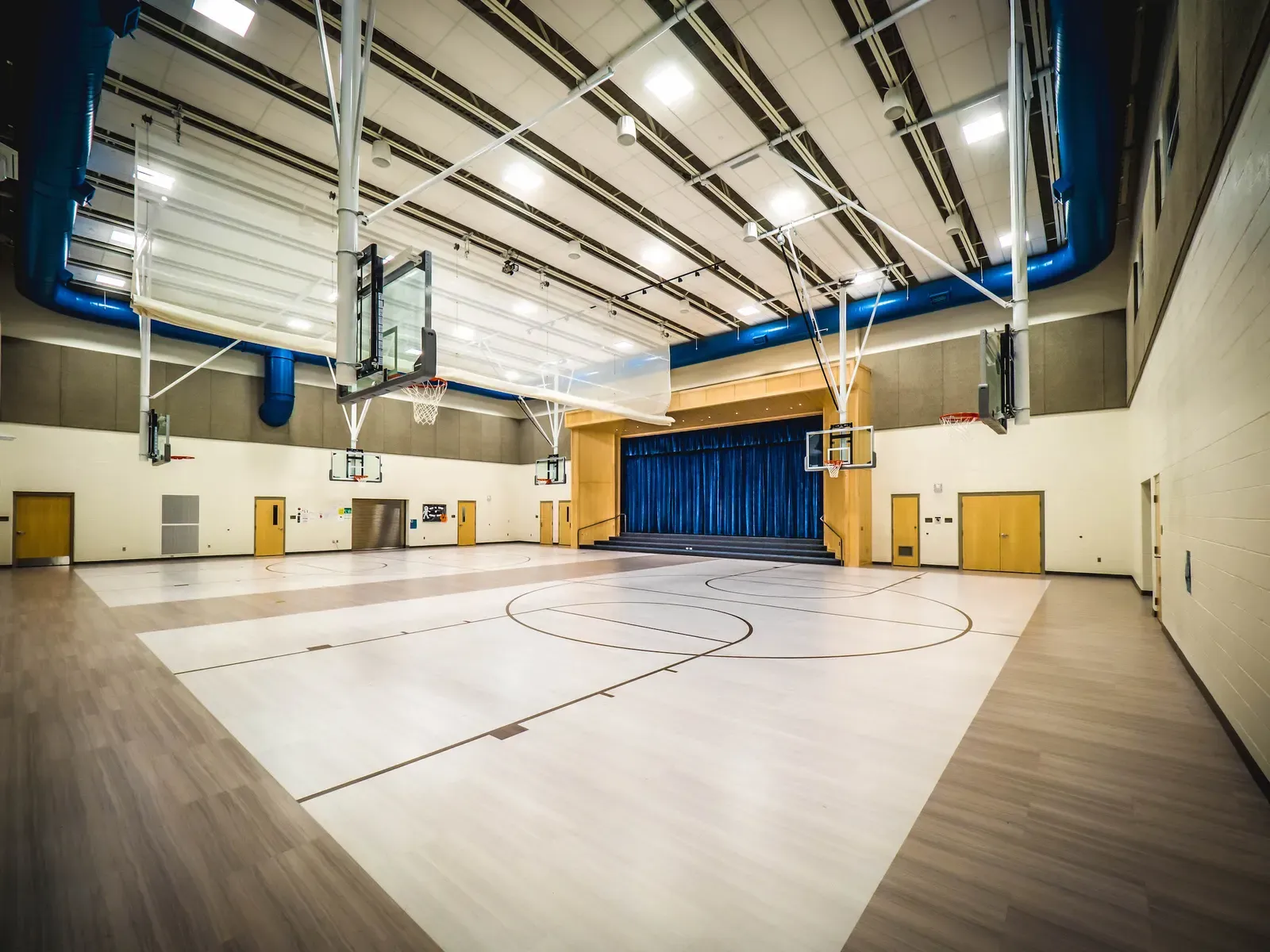 Gymnasium with a light wood floor, fold-up basketball backboards and a blue stage curtain at the far end