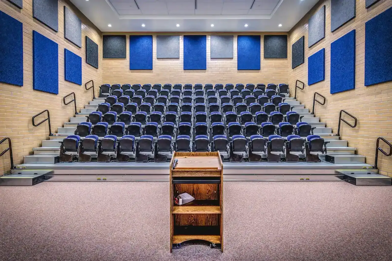 Tiered room with blue upholstered seats, blue acoustic wall panels and a wood lectern in front.