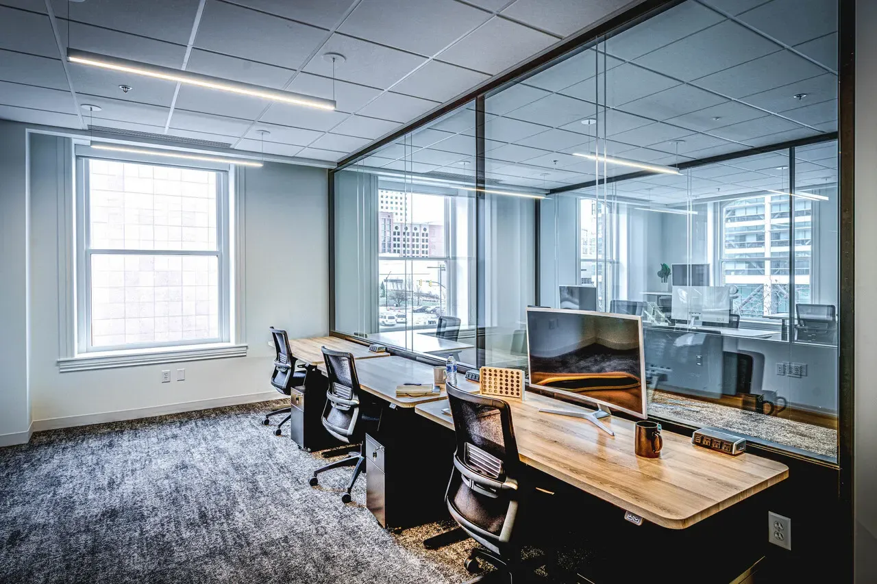 Shared office with wood-top desks, glass walls and a tall window.