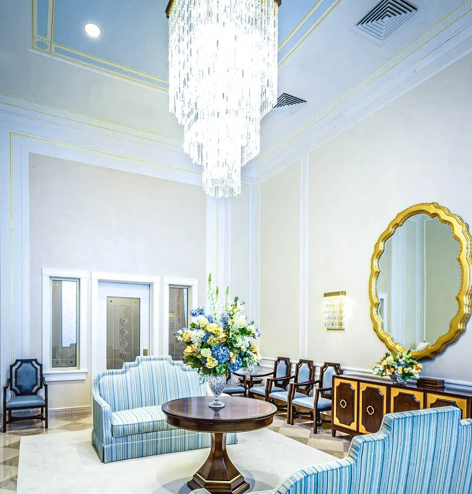 Tall sitting room with a hanging crystal chandelier, striped sofas, a gold mirror and a floral arrangement.
