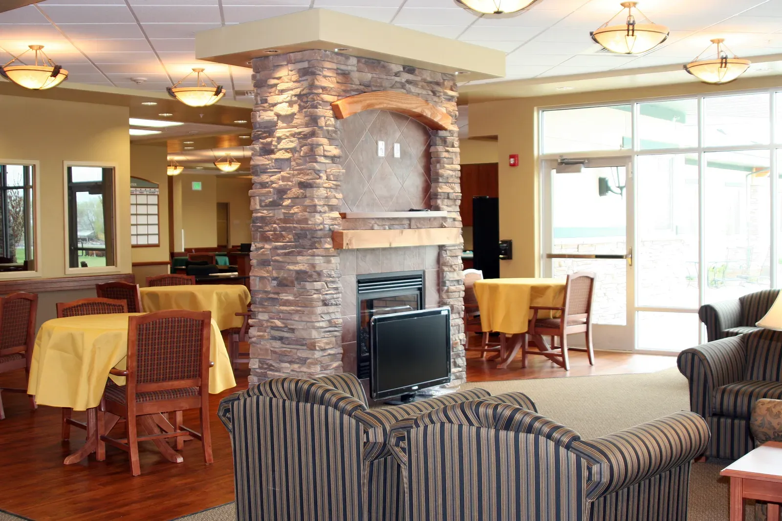 Lounge with striped armchairs facing a stone fireplace column, with dining tables and glass doors beyond.