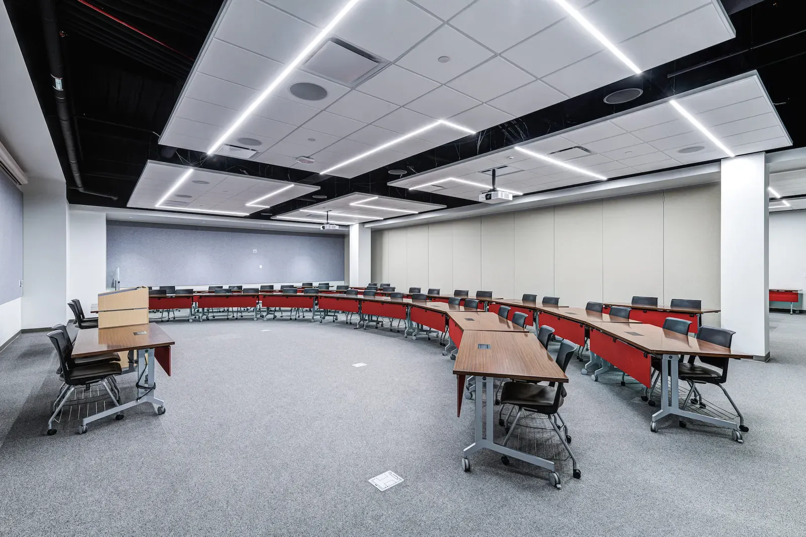 Classroom with curved rows of desks with red fronts, gray carpet and linear ceiling lights.
