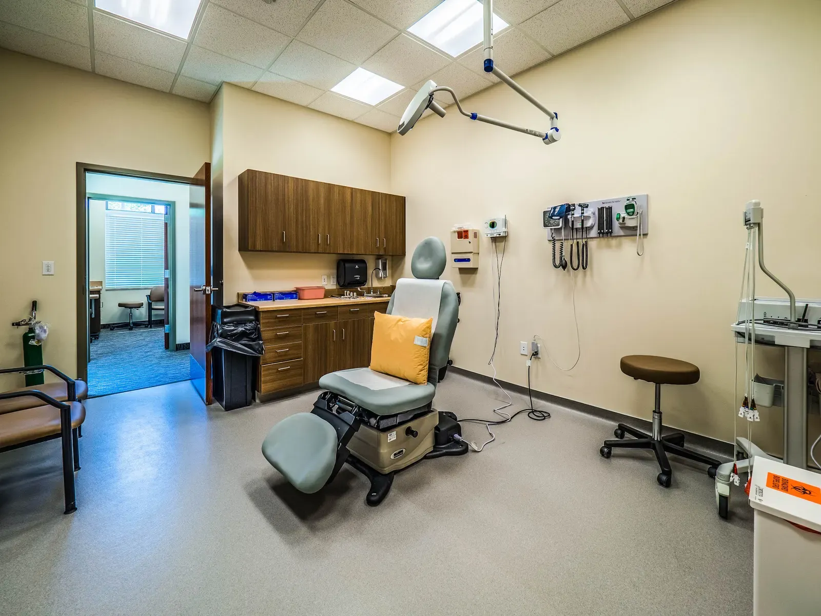 Exam room with a padded procedure chair, wall-mounted instruments, wood cabinets and a stool.