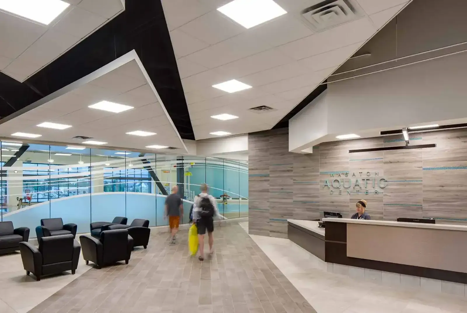 Lobby reception desk with wood-look wall panels and lounge chairs, with the pool visible through glass at left.