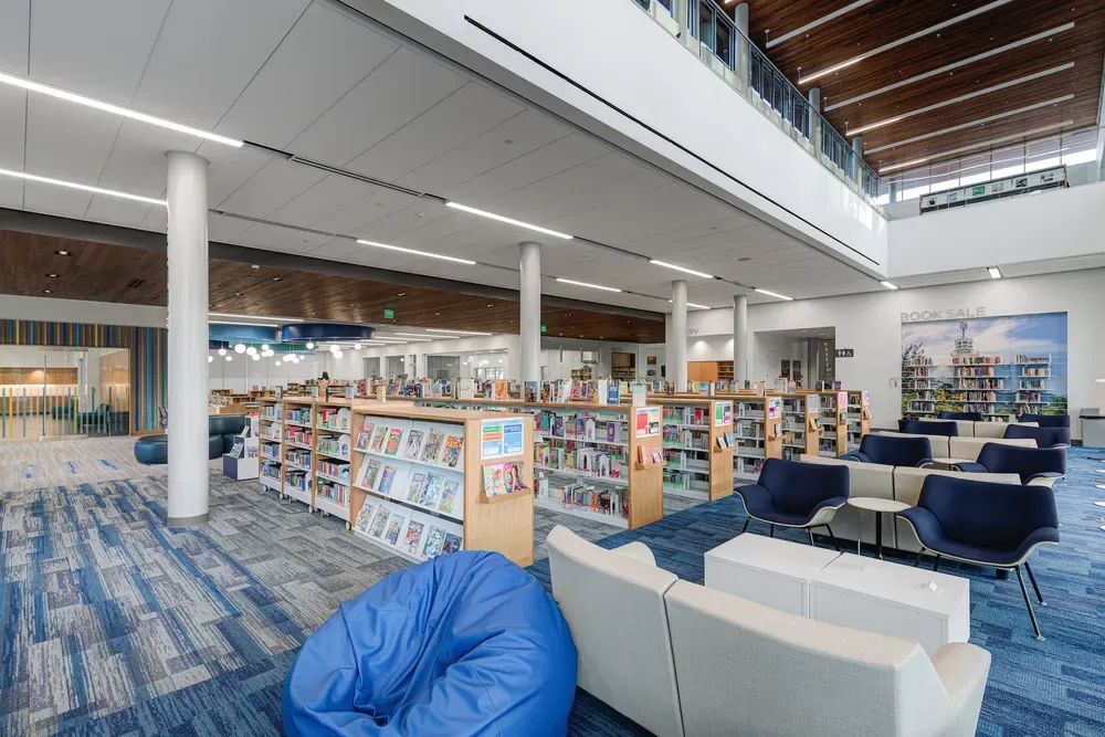 Open library stacks with lounge seating and a bean bag beneath a wood-slat ceiling, with a second-floor balcony above