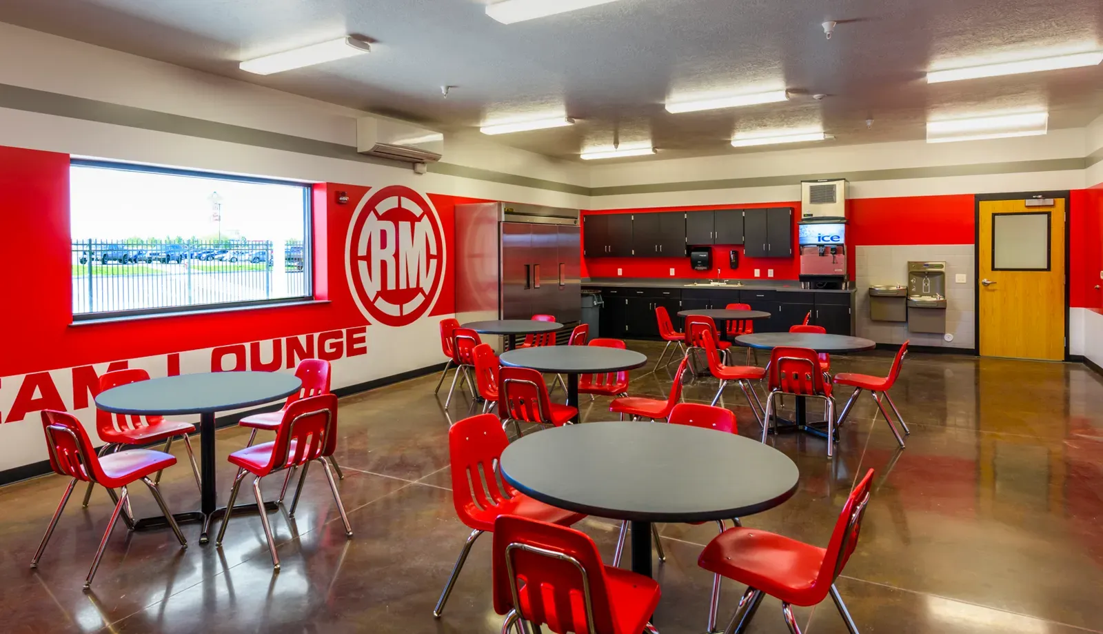 Employee lounge with red chairs, round tables, a kitchenette and a large logo on a red wall.