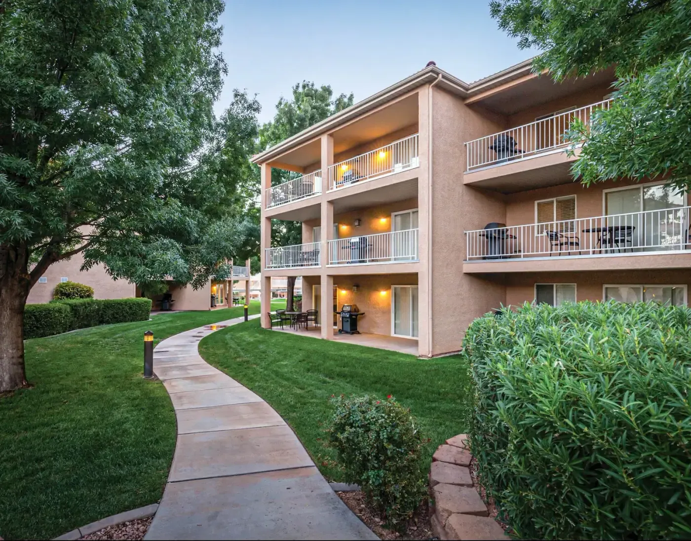 Three-story stucco building with balconies beside a curving walkway and green lawn at dusk