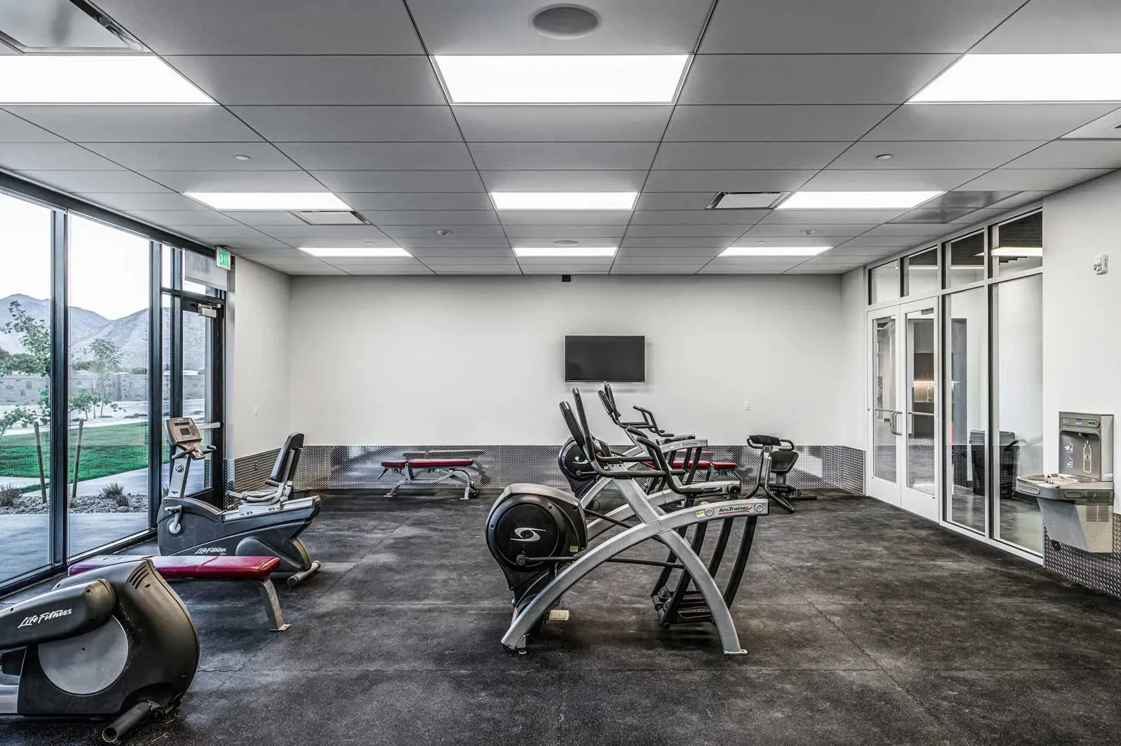 Fitness room with cardio machines and benches on a rubber floor beside a window wall.