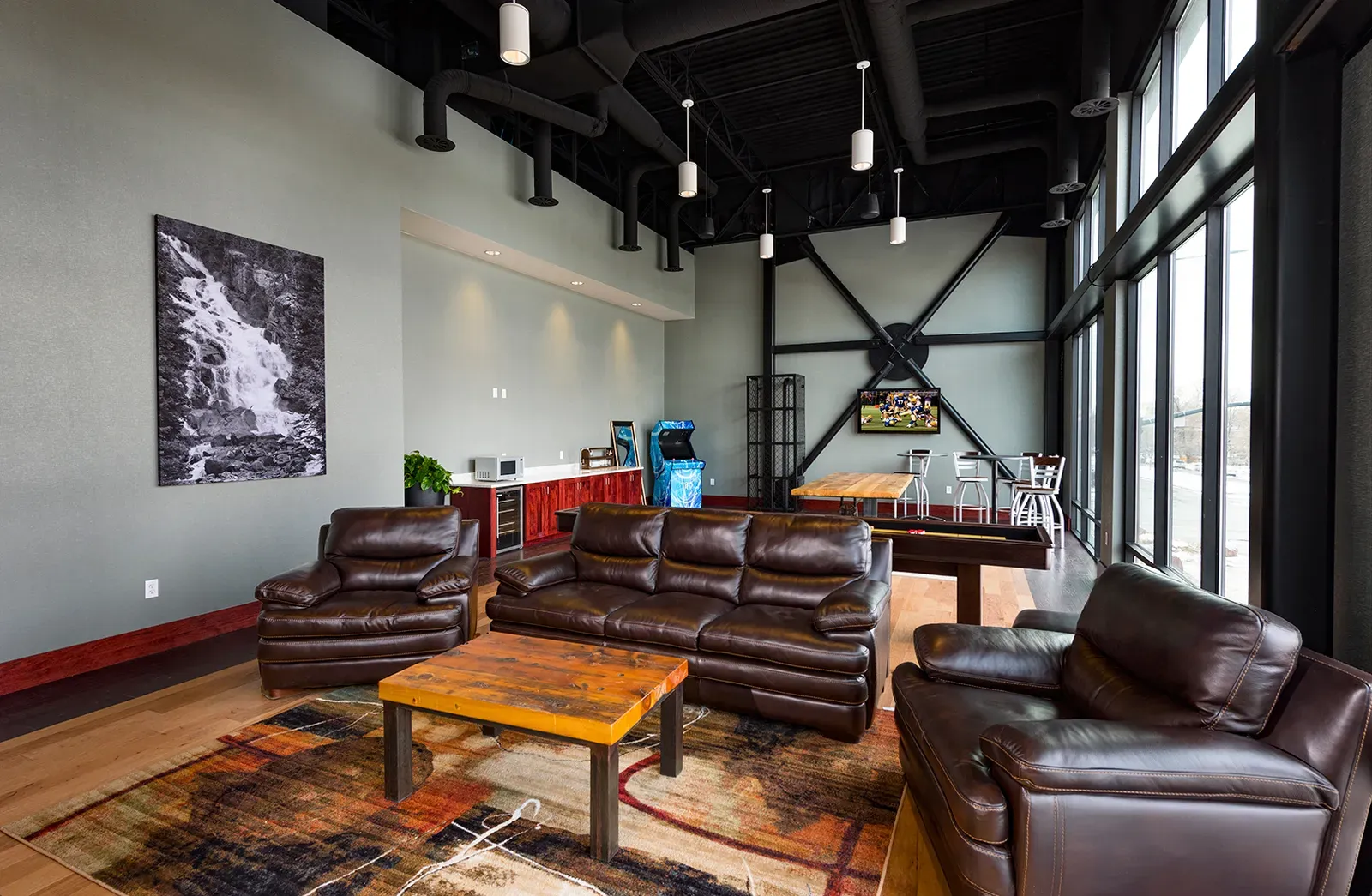 Lounge with brown leather sofas, a black-and-white waterfall photograph, an arcade game and a bar counter.