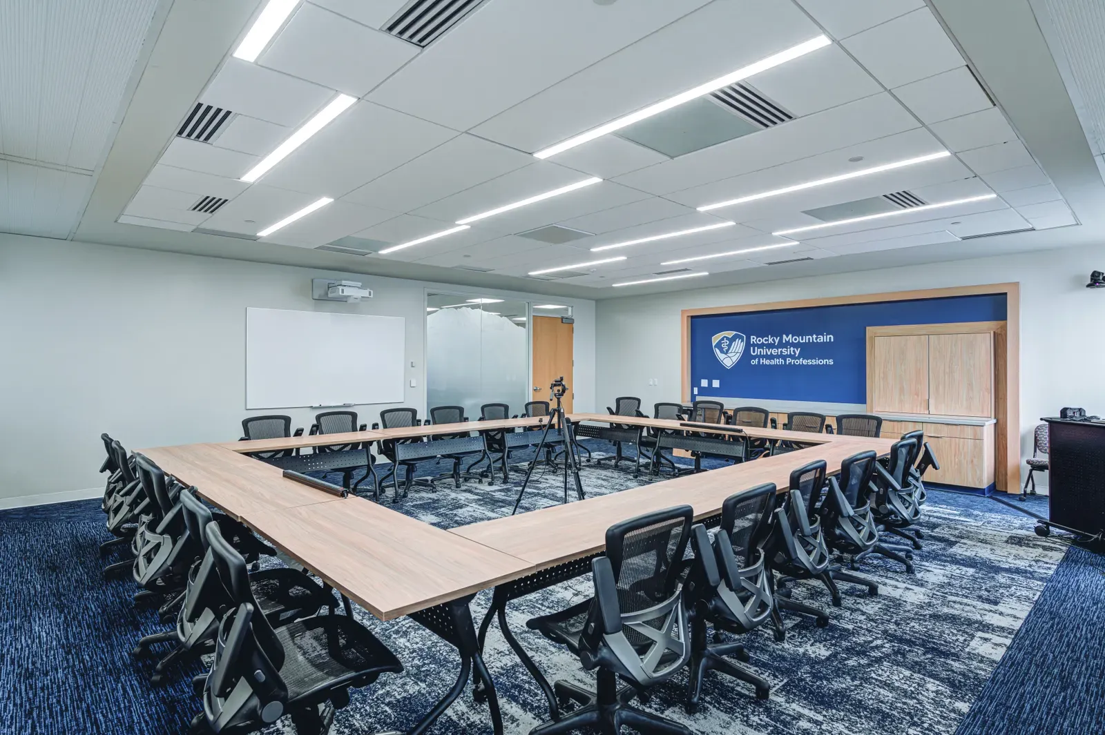 Meeting room with a large U-shaped wood table, black mesh chairs and a Rocky Mountain University sign on a blue wall.