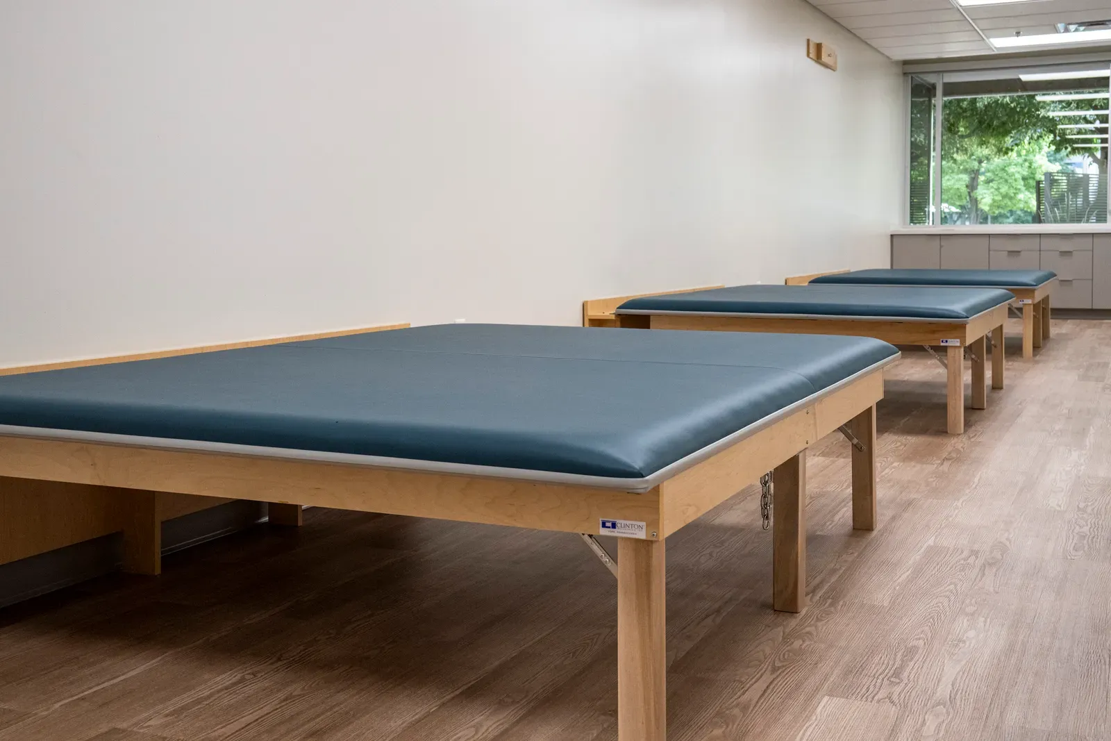 Row of padded blue treatment tables along a wall, with windows and cabinets at the far end.