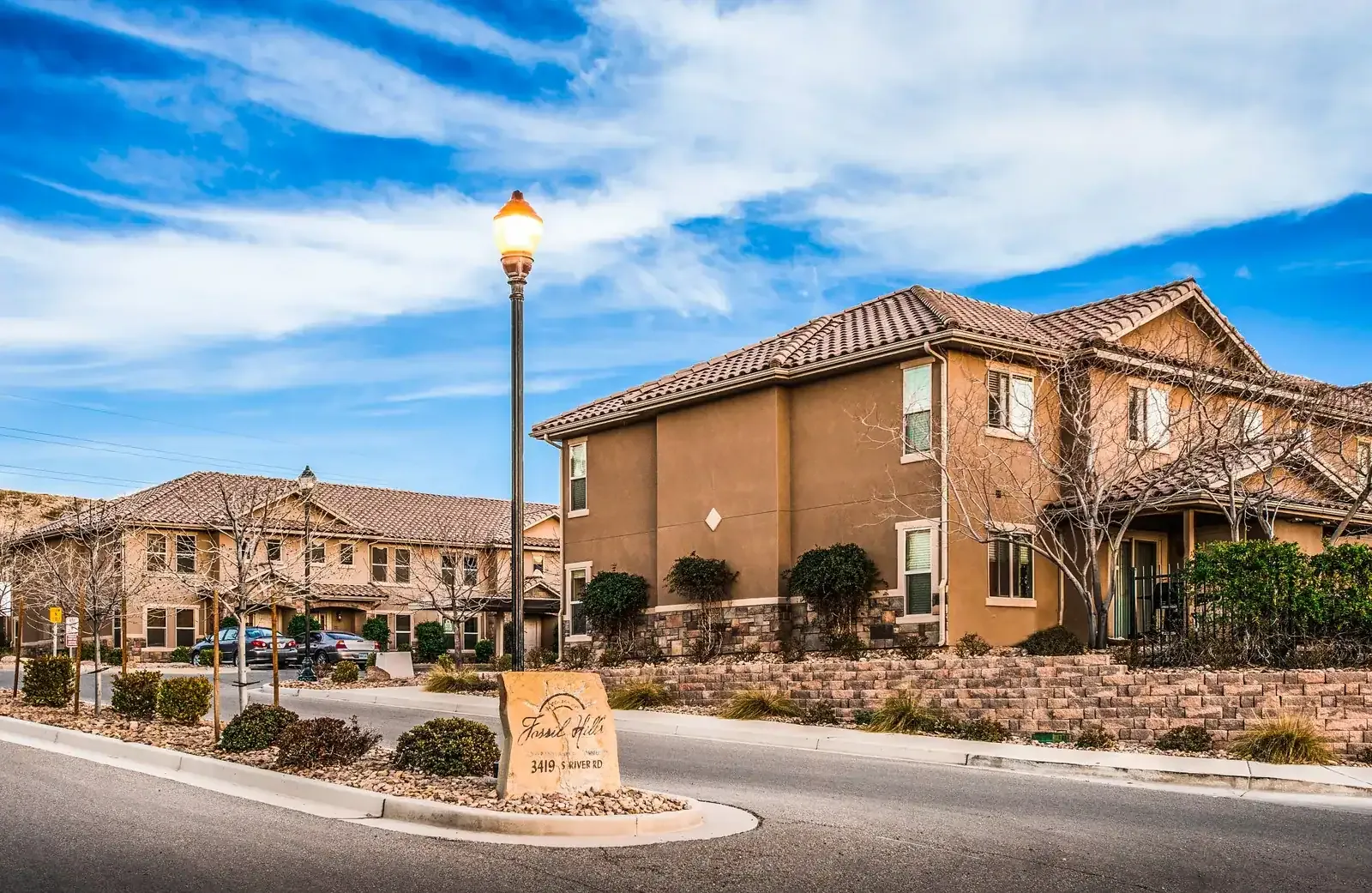 Stucco townhomes with tile roofs and a stone retaining wall along a street, with a community sign in the foreground