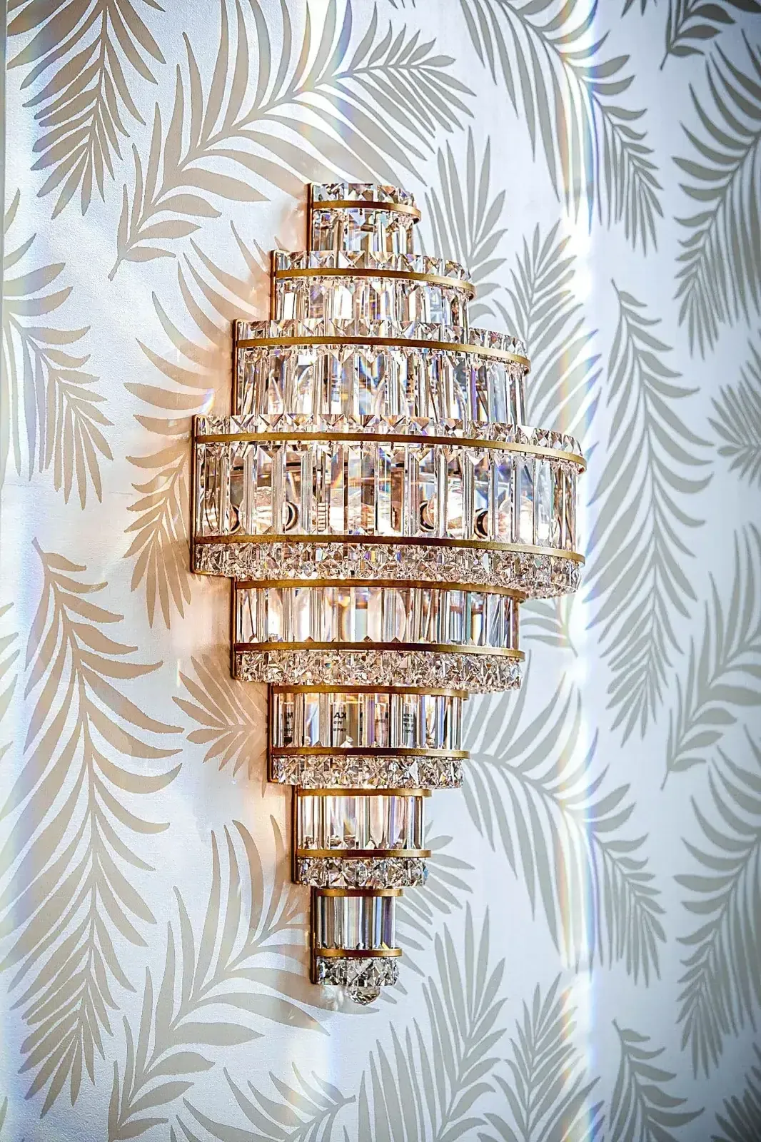 Gold and crystal wall sconce against leaf-patterned wallpaper.