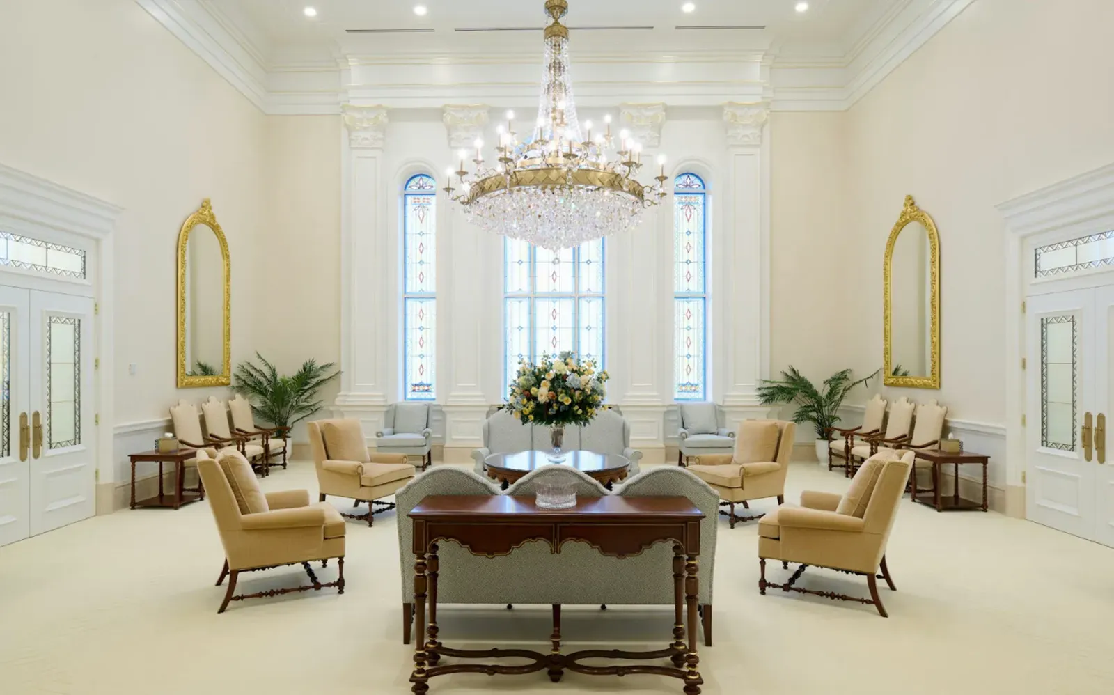 Large room with a crystal chandelier, gold-framed mirrors, arched stained-glass windows, armchairs and a table with a flower arrangement
