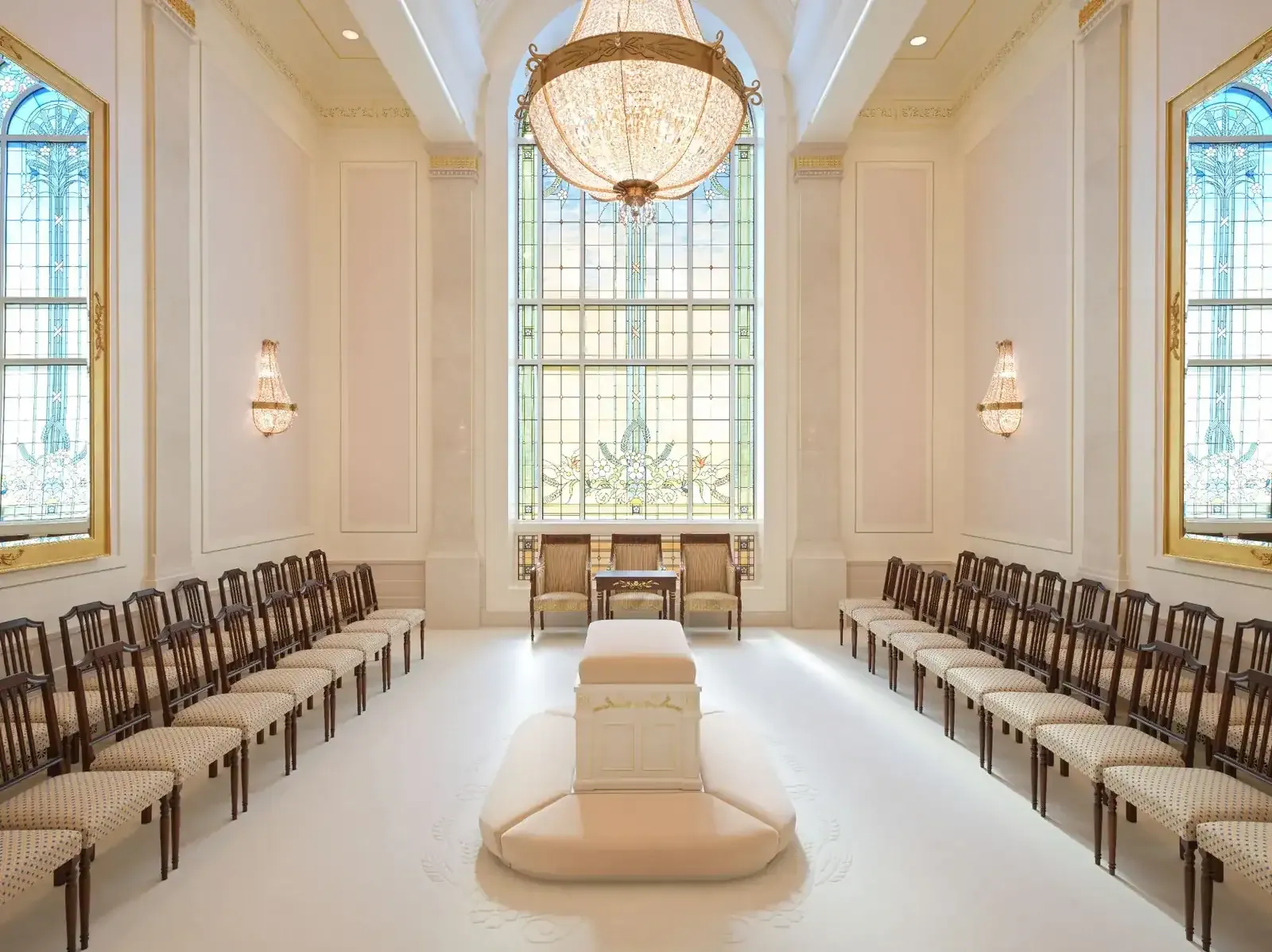 Sealing room with a central cushioned altar, rows of chairs along both walls, a crystal chandelier and a tall stained-glass window