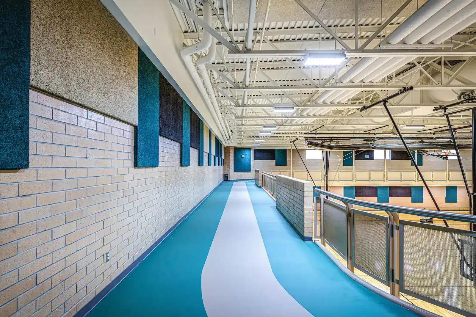 Elevated teal running track beside a brick wall with sound-absorbing panels, overlooking the gymnasium below.