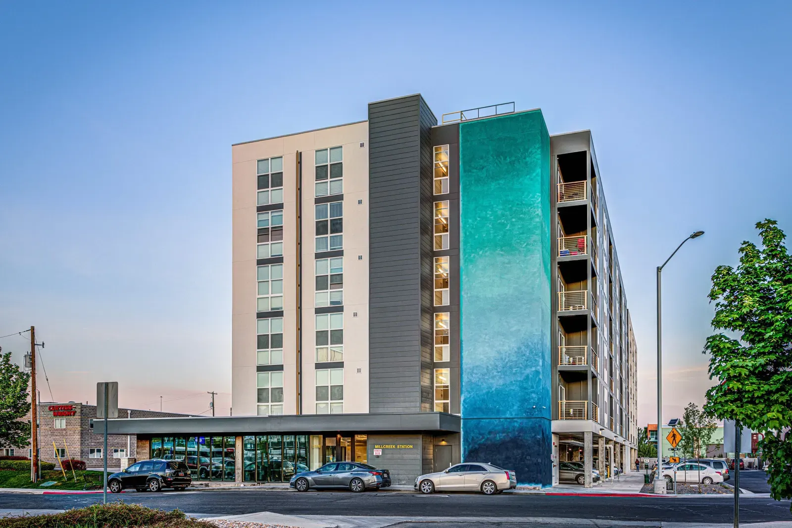 Corner of the apartment building at dusk showing the teal and blue tower, balconies and a glass ground floor.