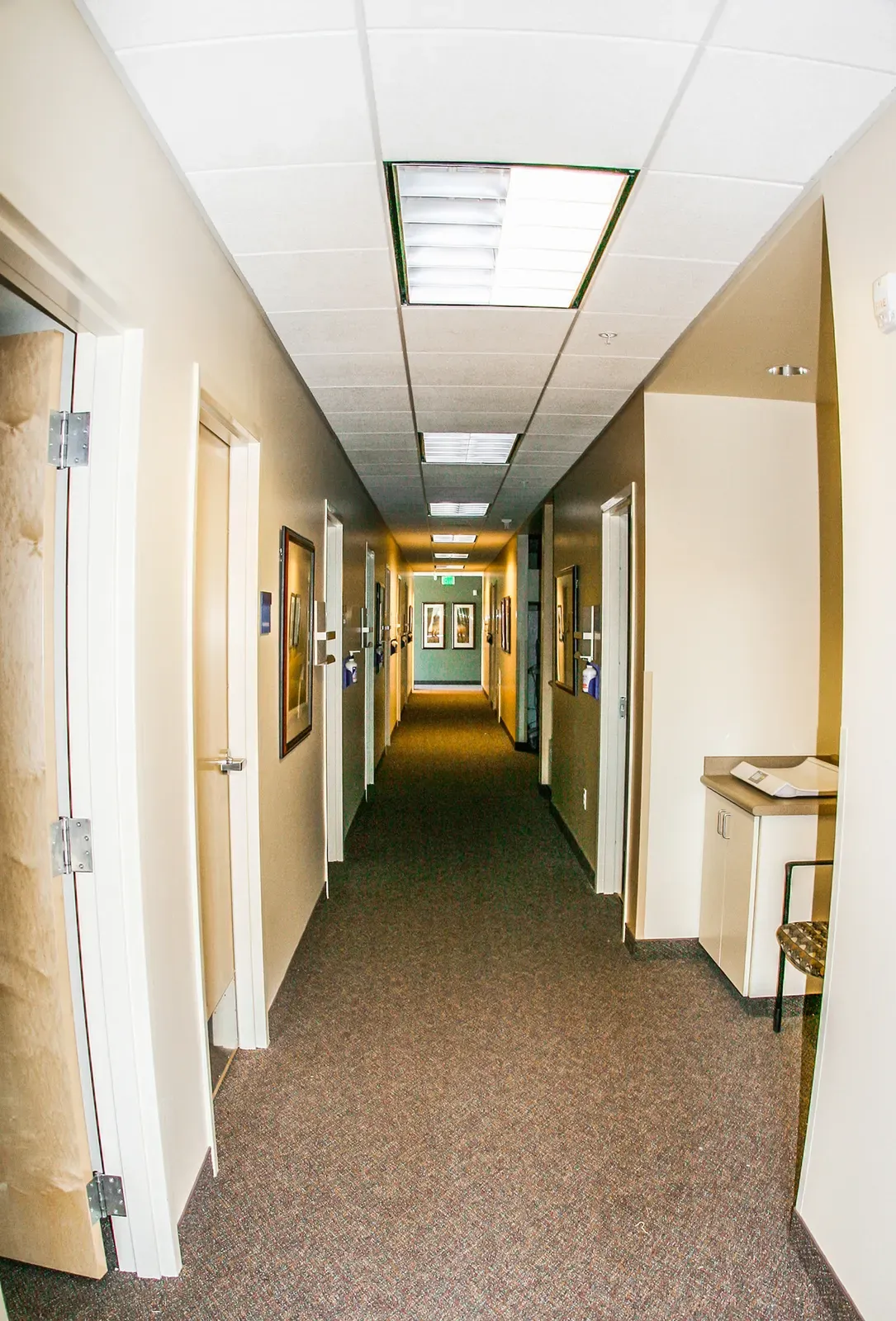 Long clinic hallway with carpet, framed art on the walls and exam room doors along both sides