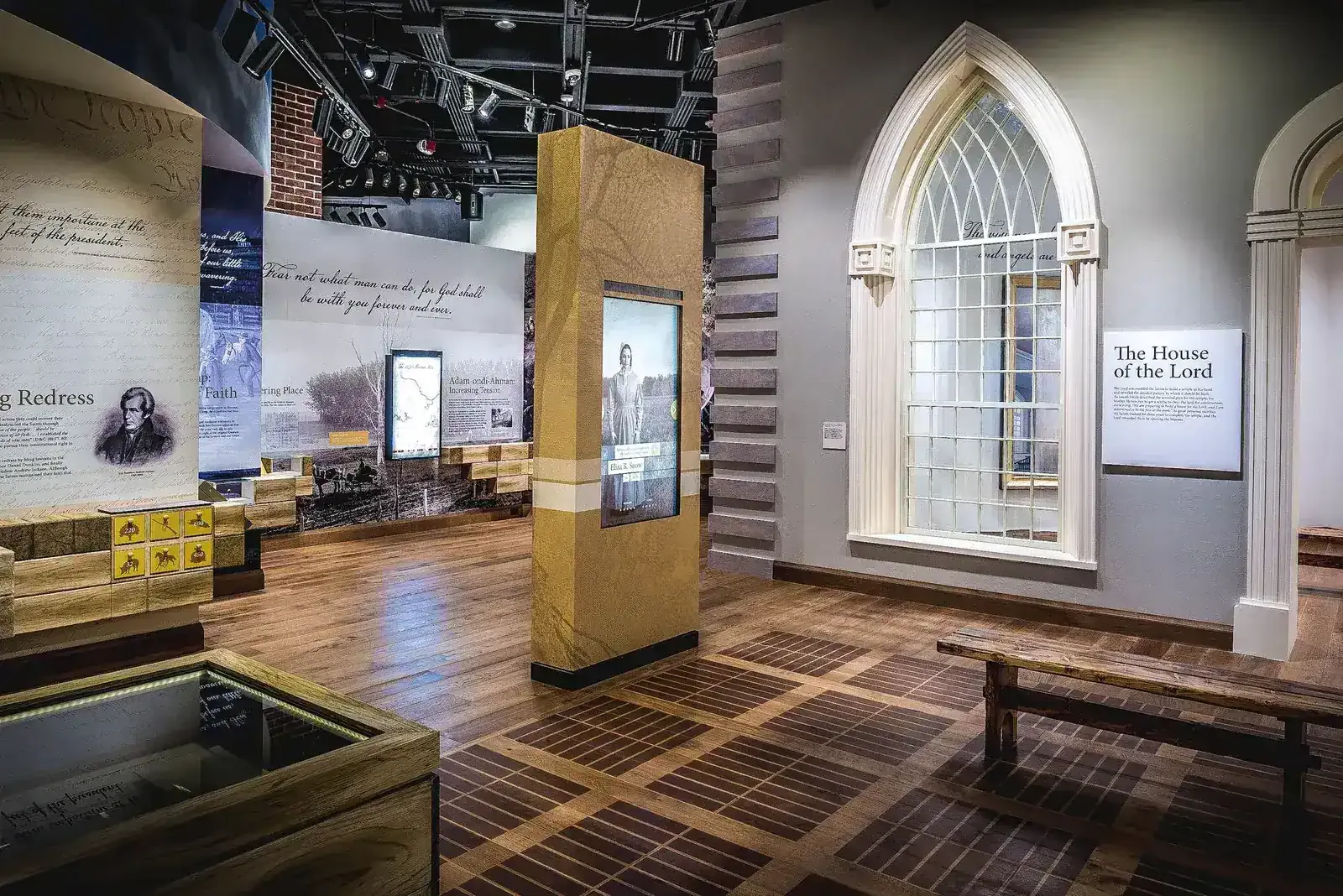 Exhibit gallery with a pointed-arch window, a patterned wood floor, and text panels on the walls