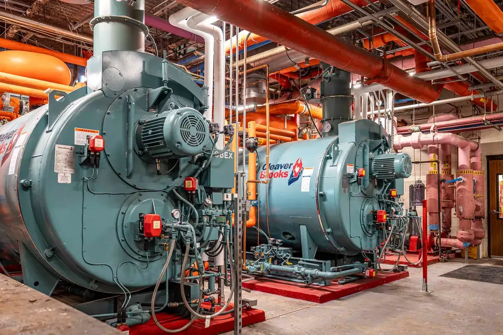 Two green boilers with orange and pink piping in the boiler room.
