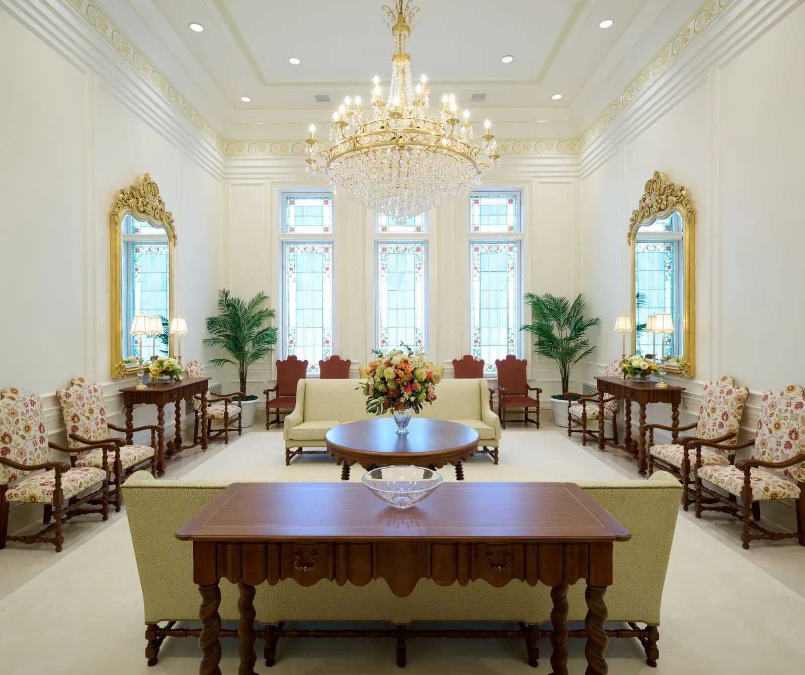 Sitting room with a crystal chandelier, tall art glass windows, gold-framed mirrors, upholstered sofas and chairs and a round wood table with flowers.