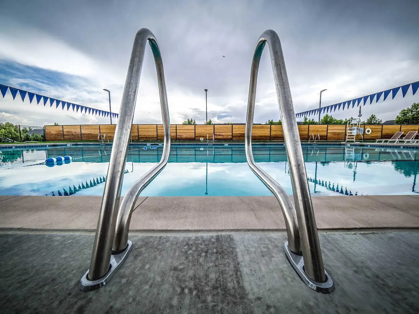 Pool ladder handrails in the foreground with the pool, banners and wood fence behind.