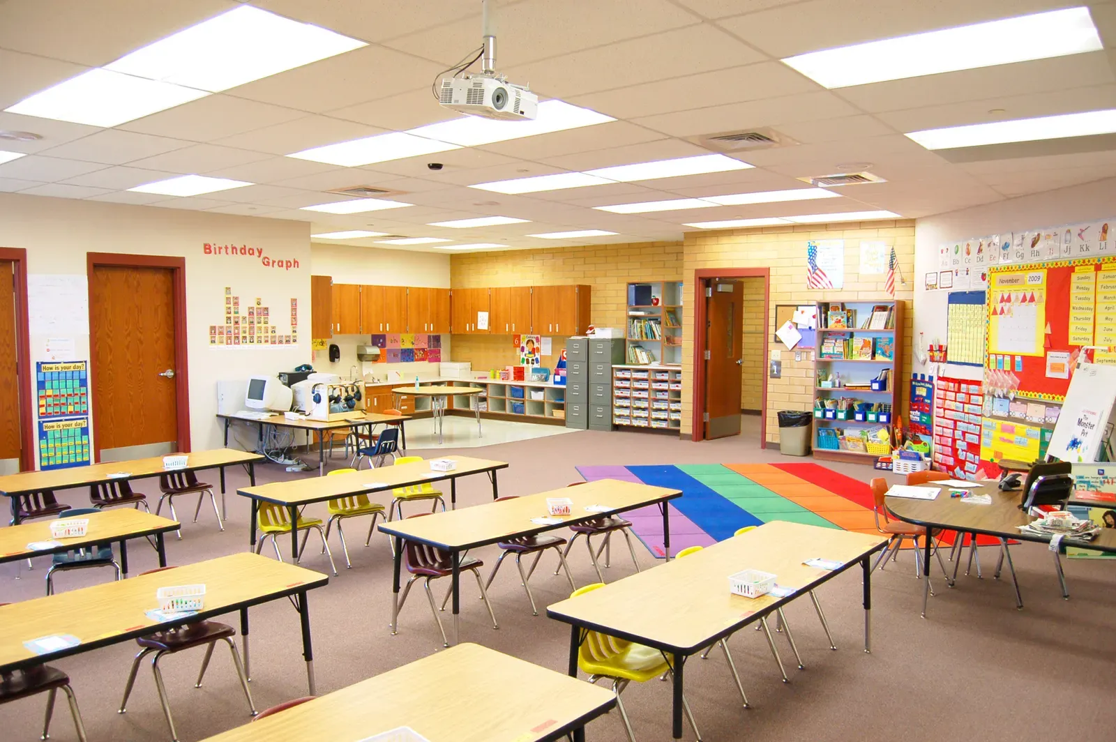 Classroom with student desks and yellow chairs, a rainbow-striped rug, bulletin boards and a ceiling-mounted projector.