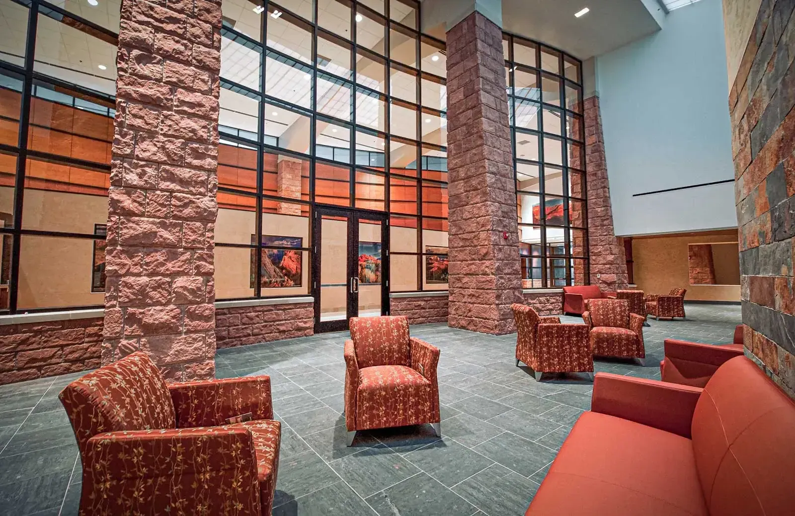 Lobby with red stone columns, a slate tile floor, orange sofas and patterned armchairs below tall windows.