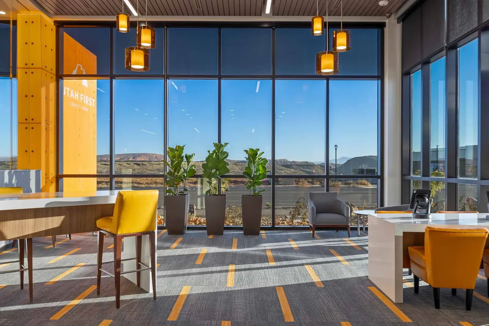 Lobby windows looking out to hills, with an orange Utah First panel, potted plants, and yellow stools.