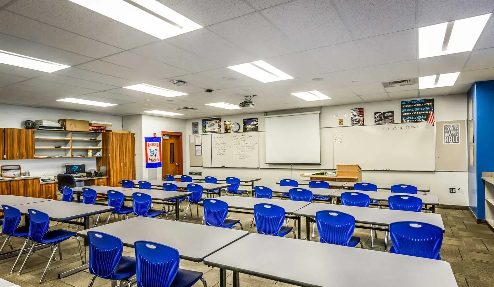 Classroom with rows of gray tables and blue chairs facing whiteboards and a projector screen
