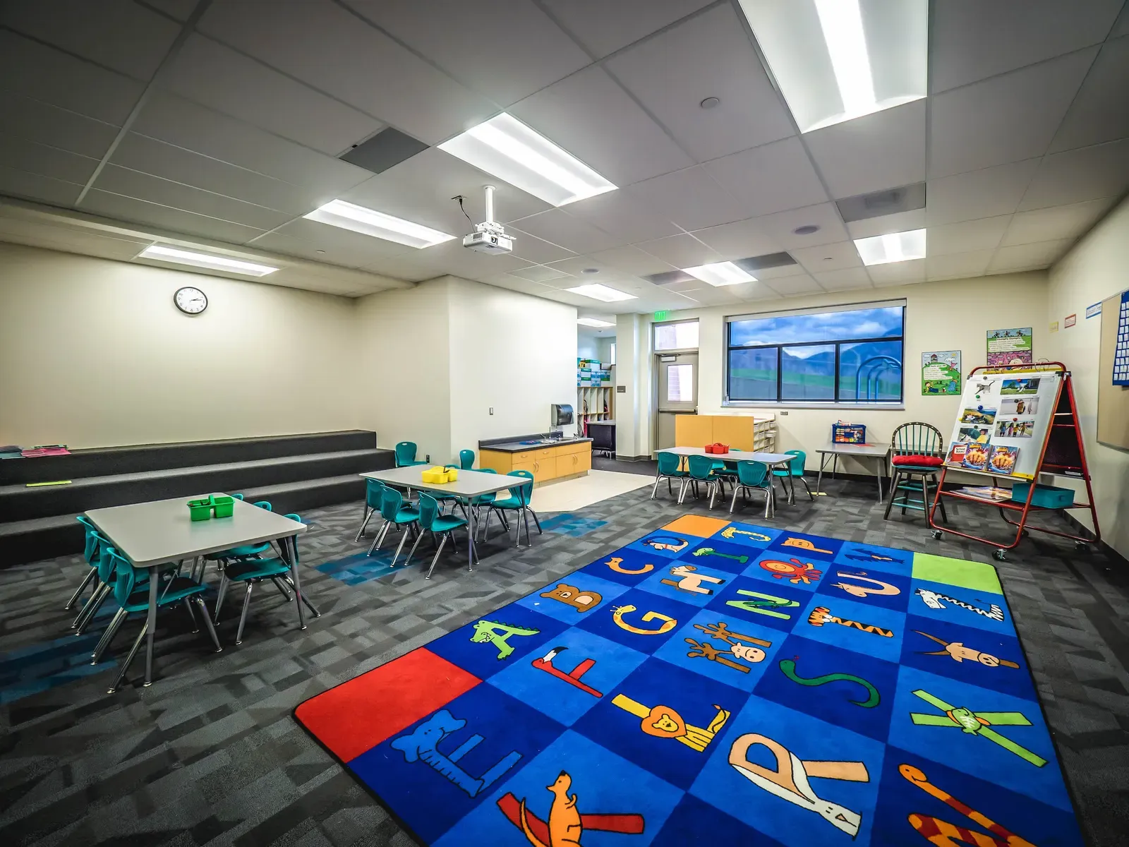 Early-grades classroom with small tables, teal chairs and an alphabet rug