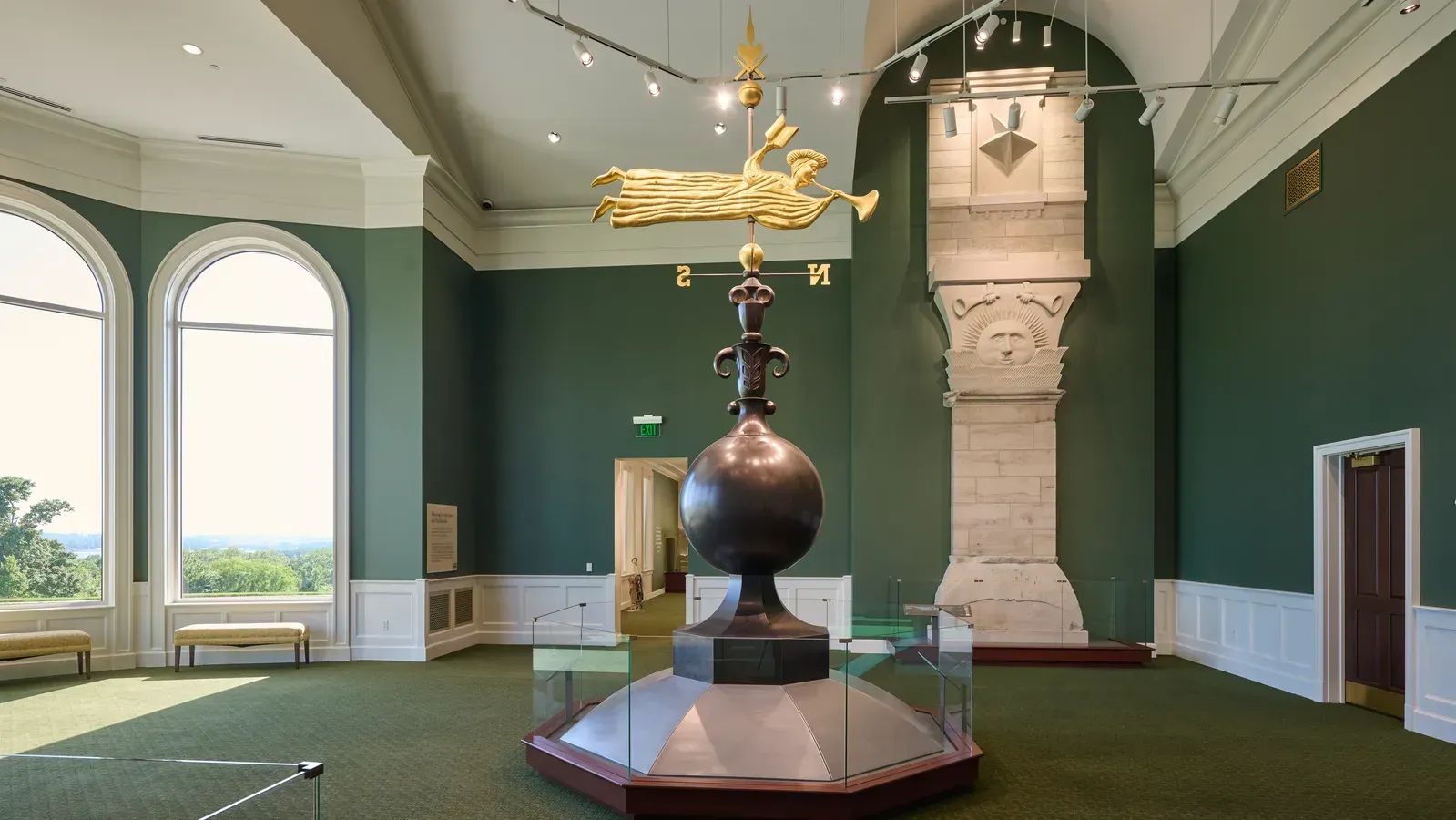 Exhibit space with a gold angel weathervane on a dark sphere in a glass case, beside a tall stone column topped with a carved sun face.