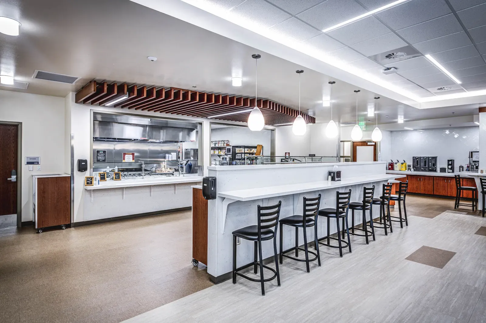 Cafeteria serving area with a counter, black bar stools and pendant lights under a wood-slat ceiling feature