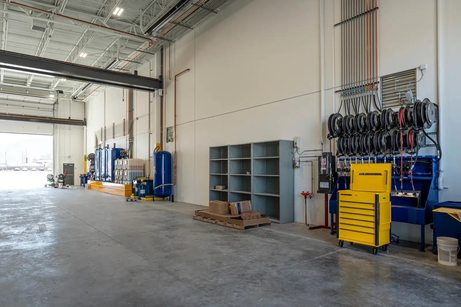 Truck shop interior with a concrete floor, an open overhead door, blue air tanks and fluid hose reels on the wall.