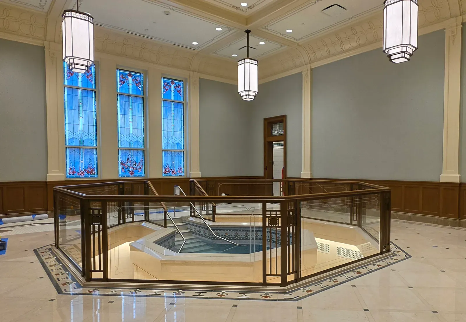 Baptismal font ringed by a glass and bronze railing on a tile floor with a mosaic border, beneath three blue art glass windows.