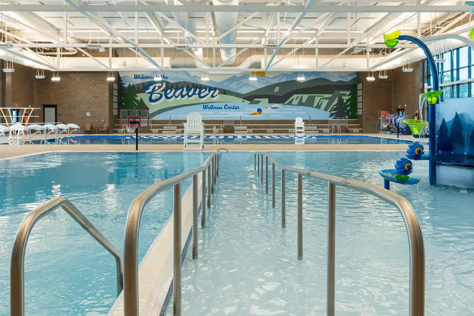 Zero-entry slope into the activity pool with stainless handrails, with the mural and a play structure beyond.