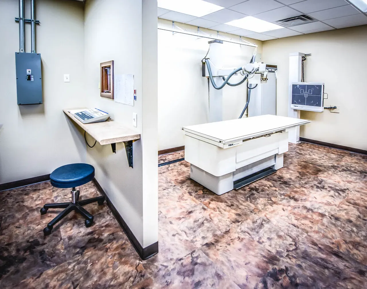 X-ray room with a radiography table, overhead tube and wall-mounted monitor.